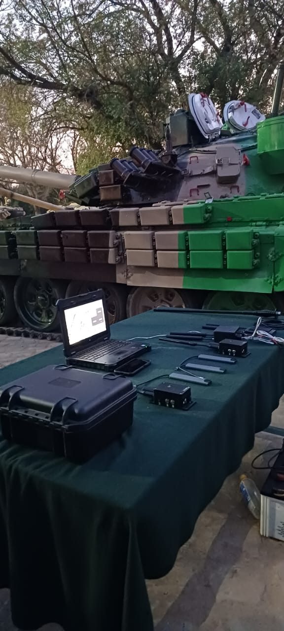 Mesh nodes, cabling and a rugged laptop laid out on a field table in front of a main battle tank