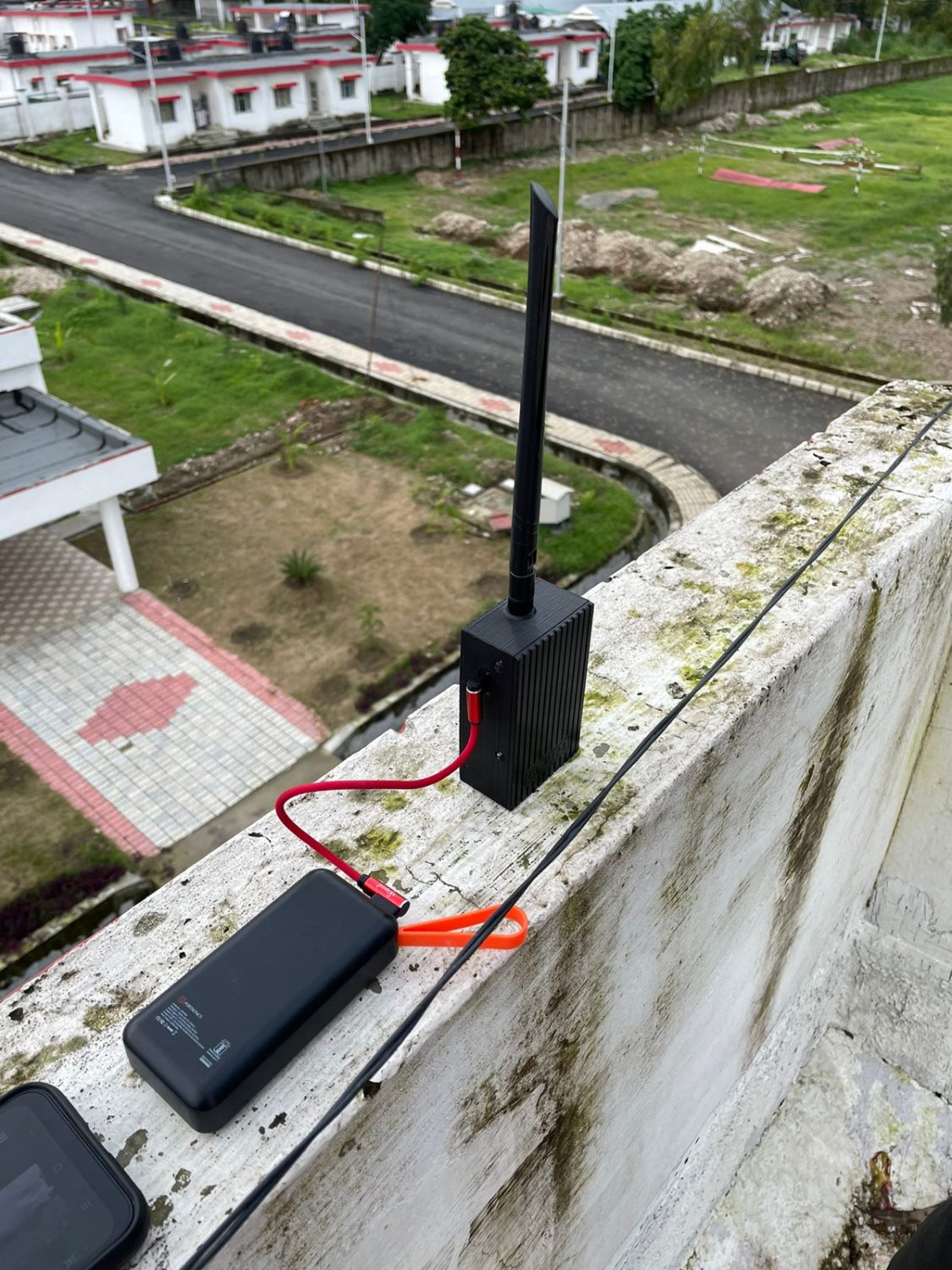 Relay node with a high-gain whip antenna clamped to a rooftop parapet, running off a portable power bank